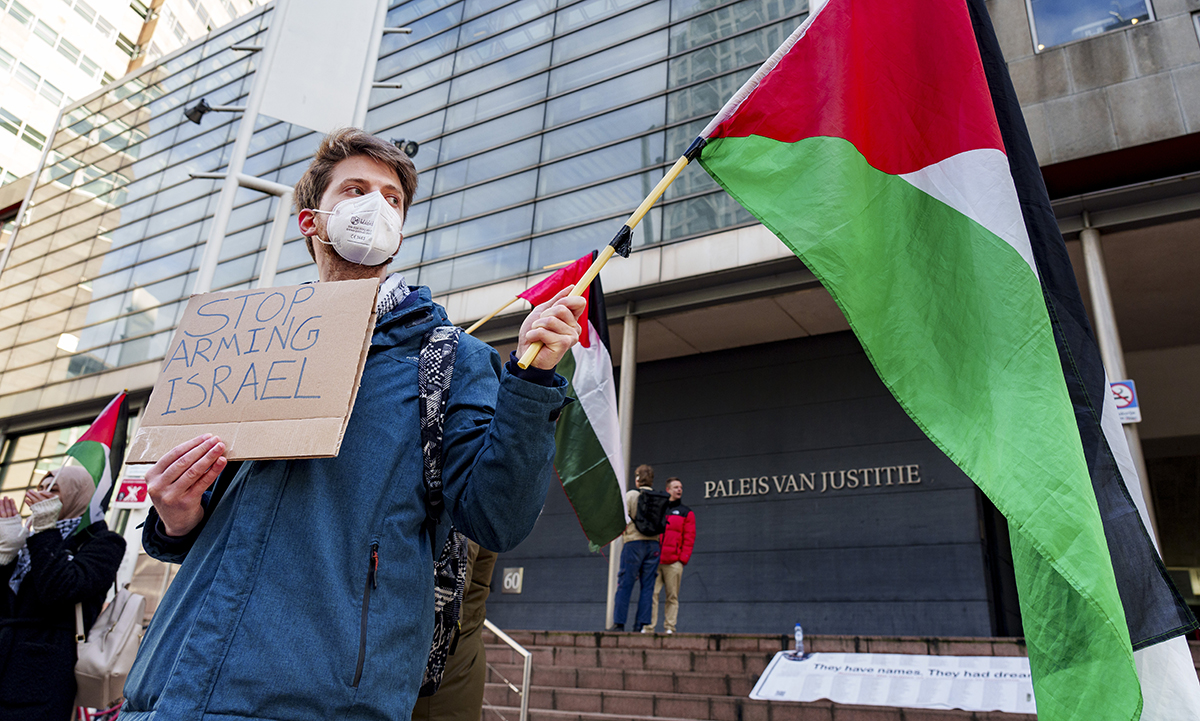 Man met een Palestijnse vlag en een bordje met 'Stop arming Israel' demonstreert voor het Paleis van Justitie